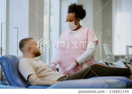 Medical professional in mask attending reclining patient in modern clinic setting with windows. Medical equipment visible in background creating professional healthcare environment 119801144