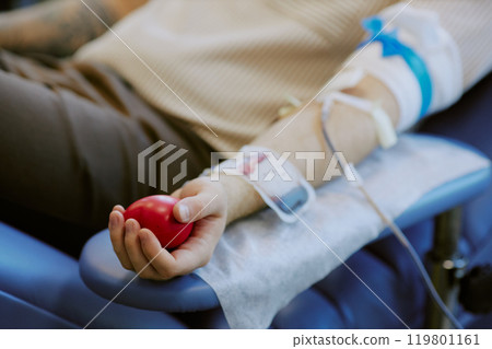 Close-up of person donating blood, holding a red squeezy ball. Arm showing IV and medical setup in a clinical setting with visible sterile procedure 119801161