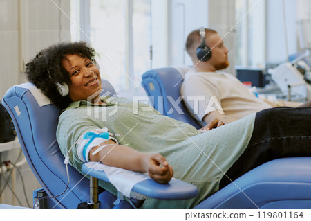 African American woman and Caucasian man donating blood while relaxing in reclining chairs. They are listening to headphones while giving blood in a bright medical facility African American woman and Caucasian man donating blood while relaxing in reclining chairs. They are listening to headphones while giving blood in a bright medical facility 119801164