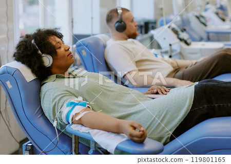 African American person donating blood while listening to music with other patients in background Donor sitting on a recliner with an IV in arm African American person donating blood while listening to music with other patients in background Donor sitting on a recliner with an IV in arm 119801165