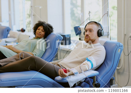 Man wearing headphones and a woman reclined in chairs bay. Engaging in donation at modern clinic while looking relaxed and comfortable 119801166