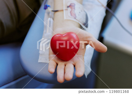 Close-up image showing person holding heart-shaped stress ball while donating blood. IV line visible on arm with background slightly blurred, emphasizing action Close-up image showing person holding heart-shaped stress ball while donating blood. IV line visible on arm with background slightly blurred, emphasizing action 119801168