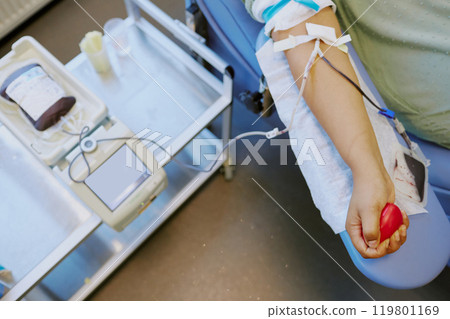 Person donating blood while sitting in medical chair, holding a stress ball in hand, with medical equipment visible around. Comprising a hospital setting with focus on blood donation process 119801169