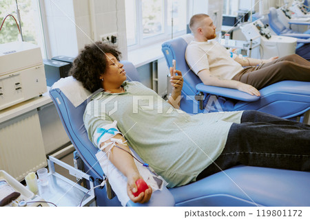 People reclined donating blood in medical facility, holding stress balls in arms Relaxing on chairs with medical equipment surrounding them, giving blood in safe and sterile environment 119801172
