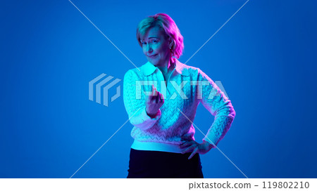 Elderly woman looking directly at camera, gesturing against blue studio background in neon light Elderly woman looking directly at camera, gesturing against blue studio background in neon light 119802210