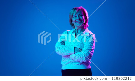 Portrait of smiling, mature, elderly woman standing with arms crossed and cheerful face against blue studio background in neon light. 119802212