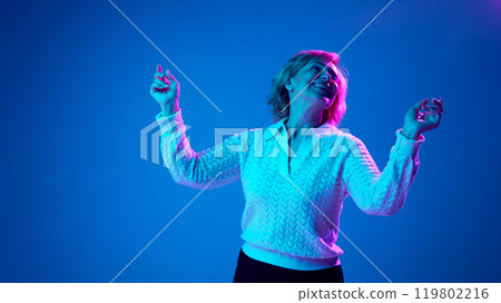 Positive, elderly woman cheerfully dancing, relaxing against blue studio background in neon light 119802216