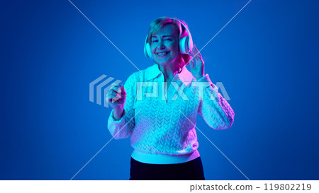 Portrait of smiling, mature, elderly woman listening to music in headphone and dancing against blue studio background in neon light Portrait of smiling, mature, elderly woman listening to music in headphone and dancing against blue studio background in neon light 119802219