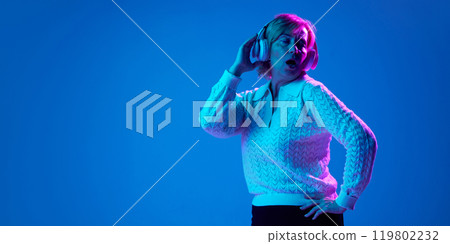 Portrait of mature, elderly woman listening to music in headphone,singing and dancing against blue studio background in neon light 119802232