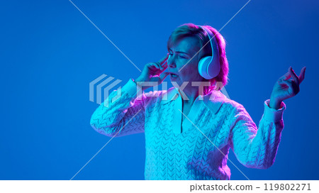 Portrait of mature, elderly woman listening to music in headphone,singing and dancing against blue studio background in neon light 119802271
