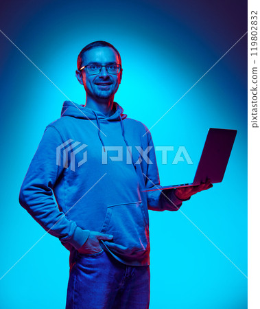 Young man, IT specialist looks confidently and smiling at camera holding laptop in neon light against gradient studio background. 119802832