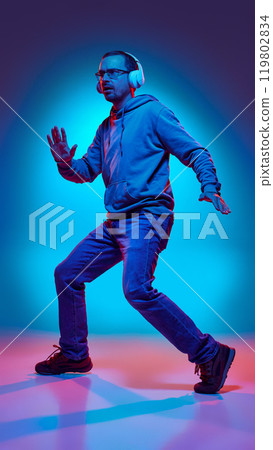 Dynamic photo of young active and funny man dancing listening music in headphones in neon light against gradient studio background. 119802834