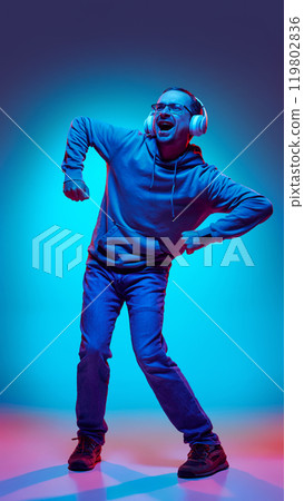 Casual dressed young man, dancing energetically listening music in headphones in neon light against gradient studio background. Casual dressed young man, dancing energetically listening music in headphones in neon light against gradient studio background. 119802836