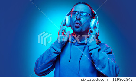 Young man listening music and sig with joy in neon light against gradient studio background. Hobby. Young man listening music and sig with joy in neon light against gradient studio background. Hobby. 119802842