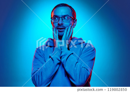 Portrait of young man dressed casual hoodie and holding hands near mouth as is he scared in neon light against gradient studio background. 119802850