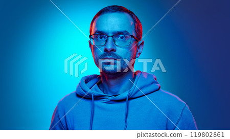 Portrait of young man in glasses dressed hoodie looking at camera with calm in neon light against gradient studio background. 119802861