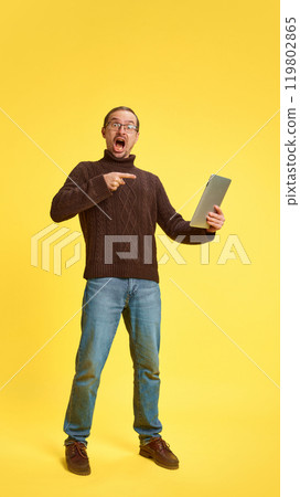 Shocked young man dressed casual attire pointing to tablet announcing sales, discounts against yellow studio background. 119802865