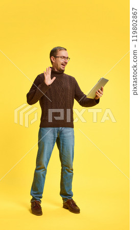 Young man dressed casual attire with tablet waving hello while holding meeting online against yellow studio background. Young man dressed casual attire with tablet waving hello while holding meeting online against yellow studio background. 119802867
