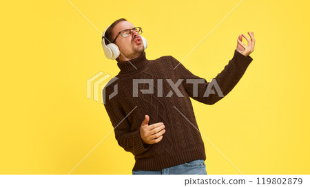 Young man in winter sweater listening music in headphones and dancing as if he playing guitar. against yellow studio background. Young man in winter sweater listening music in headphones and dancing as if he playing guitar. against yellow studio background. 119802879