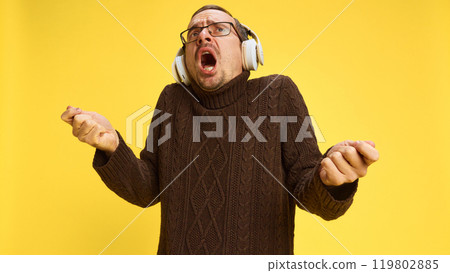 Young man in glasses with headphones, showing joyfulness while listening music in headphones and singing against yellow background. 119802885