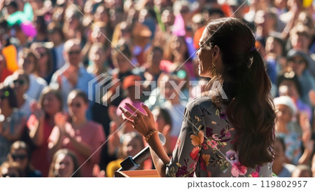 A female leader delivers a passionate speech to a diverse, enthusiastic crowd that cheers her visionary message. 119802957