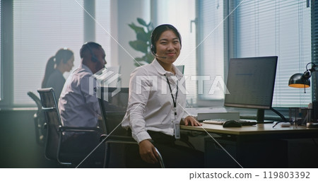 Asian female helpdesk specialist talks on headset, uses computer, looks at camera 119803392