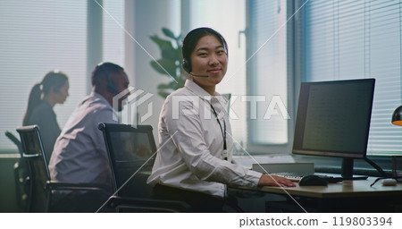 Asian female technical support operator wearing headset looks at camera Asian female technical support operator wearing headset looks at camera 119803394