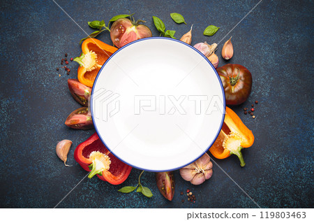 Empty white plate surrounded by fresh cooking ingredients and vegetables on dark blue background, space for text 119803463