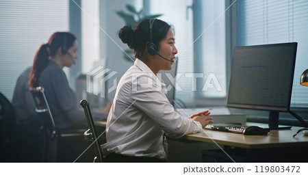 Asian female helpdesk specialist in headset consults client via call, uses computer Asian female helpdesk specialist in headset consults client via call, uses computer 119803472