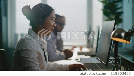 Asian technical support specialist sitting down at computer, putting on headset 119803493