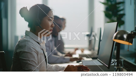 Asian technical support specialist sitting down at computer, putting on headset 119803494