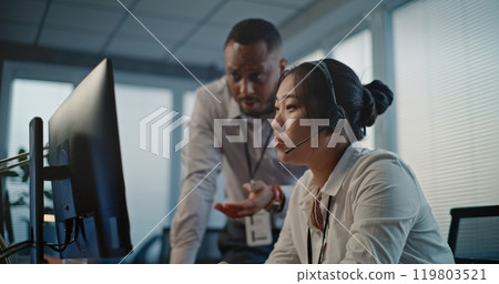 Asian helpdesk specialist in headset uses computer, talks to African American colleague Asian helpdesk specialist in headset uses computer, talks to African American colleague 119803521