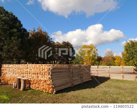 Stacked wooden planks lie on the grass against a backdrop of green and yellow trees and a blue sky with puffy clouds Stacked wooden planks lie on the grass against a backdrop of green and yellow trees and a blue sky with puffy clouds 119803522