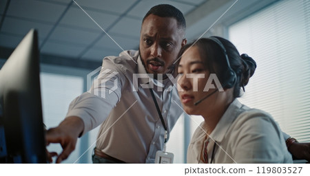Asian technical support operator in headset uses computer, talks to African American colleague Asian technical support operator in headset uses computer, talks to African American colleague 119803527