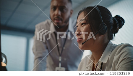 Call center office: African American technical support specialist helps Asian colleague in headset Call center office: African American technical support specialist helps Asian colleague in headset 119803529