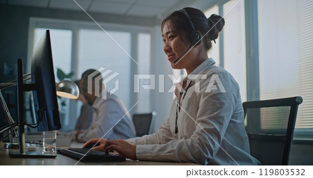 Call center office: Friendly Asian female hotline operator talking on headset, using computer 119803532