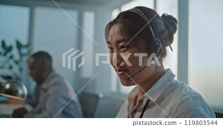 Friendly Asian female hotline specialist talks on headset, provides customer service 119803544