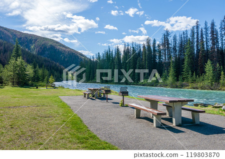 A picnic table and benches on the green meadow next to a river. Mountain and forest in the background. Maligne River in Jasper National Park, Alberta, Canada. Canadian Rockies. A picnic table and benches on the green meadow next to a river. Mountain and forest in the background. Maligne River in Jasper National Park, Alberta, Canada. Canadian Rockies. 119803870