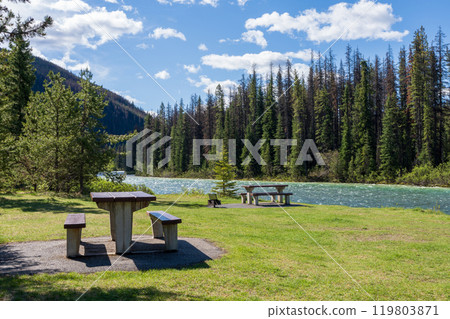 A picnic table and benches on the green meadow next to a river. Mountain and forest in the background. Maligne River in Jasper National Park, Alberta, Canada. Canadian Rockies. A picnic table and benches on the green meadow next to a river. Mountain and forest in the background. Maligne River in Jasper National Park, Alberta, Canada. Canadian Rockies. 119803871