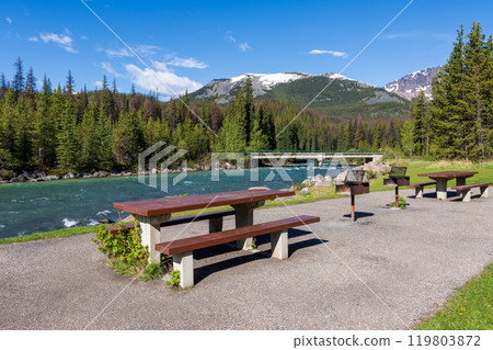 A picnic table and benches on the green meadow next to a river. Mountain and forest in the background. Maligne River in Jasper National Park, Alberta, Canada. Canadian Rockies. A picnic table and benches on the green meadow next to a river. Mountain and forest in the background. Maligne River in Jasper National Park, Alberta, Canada. Canadian Rockies. 119803872