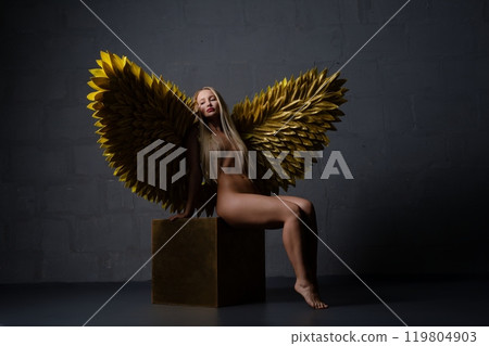 A figure with golden feathered wings poses gracefully in a dimly lit space A figure with golden feathered wings poses gracefully in a dimly lit space 119804903