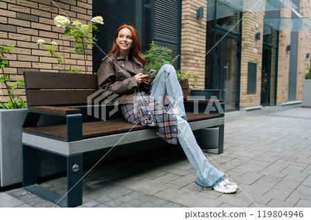 Pretty redhead female browsing smartphone, enjoying beautiful day in city. Happy red-haired young woman in leather jacket sitting on bench in city, holding using mobile phone outdoors. Pretty redhead female browsing smartphone, enjoying beautiful day in city. Happy red-haired young woman in leather jacket sitting on bench in city, holding using mobile phone outdoors. 119804946