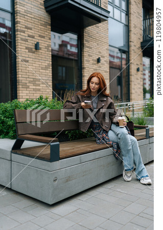 Vertical shot of happy woman with red hair enjoying coffee break sitting on bench, smiling and checking phone, embracing urban life. Cheerful redhead female using smartphone holding coffee outdoors. Vertical shot of happy woman with red hair enjoying coffee break sitting on bench, smiling and checking phone, embracing urban life. Cheerful redhead female using smartphone holding coffee outdoors. 119804957