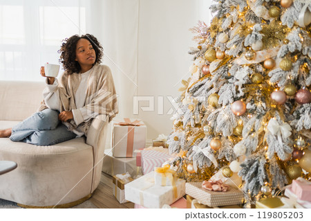 Merry Christmas. African American woman with cup of hot drink coffee tea sitting near Christmas tree at home. Girl in living room with Christmas tree. Christmas hygge holidays at home Merry Christmas. African American woman with cup of hot drink coffee tea sitting near Christmas tree at home. Girl in living room with Christmas tree. Christmas hygge holidays at home 119805203