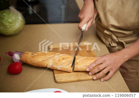 Person slicing fresh baguette on wooden cutting board in kitchen 119806057