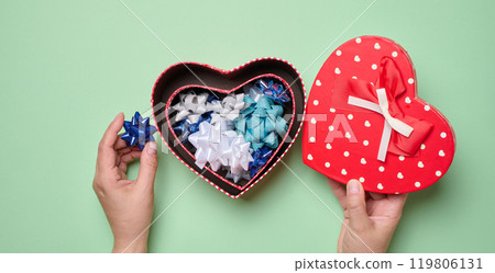 Female hands holding heart shaped gift box and bows on green background, top view Female hands holding heart shaped gift box and bows on green background, top view 119806131