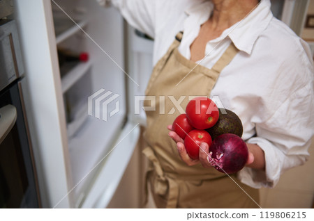Woman in apron holding fresh ingredients near open fridge 119806215
