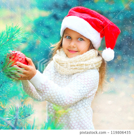 Christmas smiling child little girl in santa red hat holding ball near a branch tree Christmas smiling child little girl in santa red hat holding ball near a branch tree 119806435