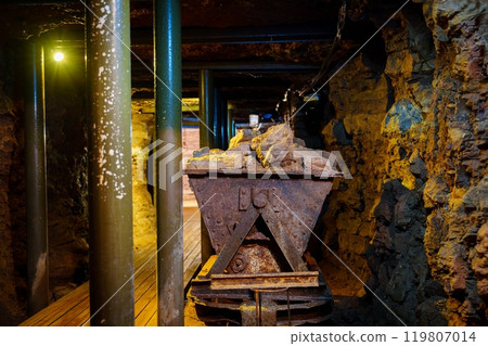 Grungy Rusty Mine Cart Sits on Old Wooden Tracks in a Dark Mine Shaft 119807014