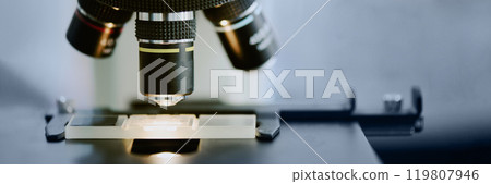Close-up of a laboratory sample being examined under a microscope for scientific research. The sample is precisely positioned under the objective lens Close-up of a laboratory sample being examined under a microscope for scientific research. The sample is precisely positioned under the objective lens 119807946